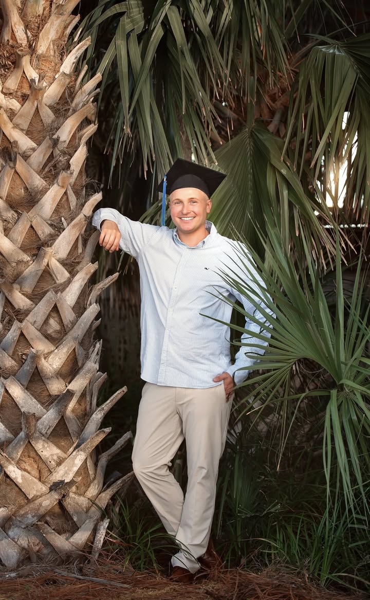 A graduate in a cap leaning against a palm tree at sunset